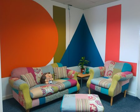 A corner of the Stronger Together Hub in the DIAL office. A colourful, patchwork style sofa set consisting of a 2 seater sofa, an arm chair and a footstool frame the photo, matching the DIAL colours almost perfectly. There is also a side table with some plants and our unofficial mascot of teddy hedgehog on the sofa. Behind the sofas, on the wall, is the new DIAL mural wall, which is a recreation of our logo.